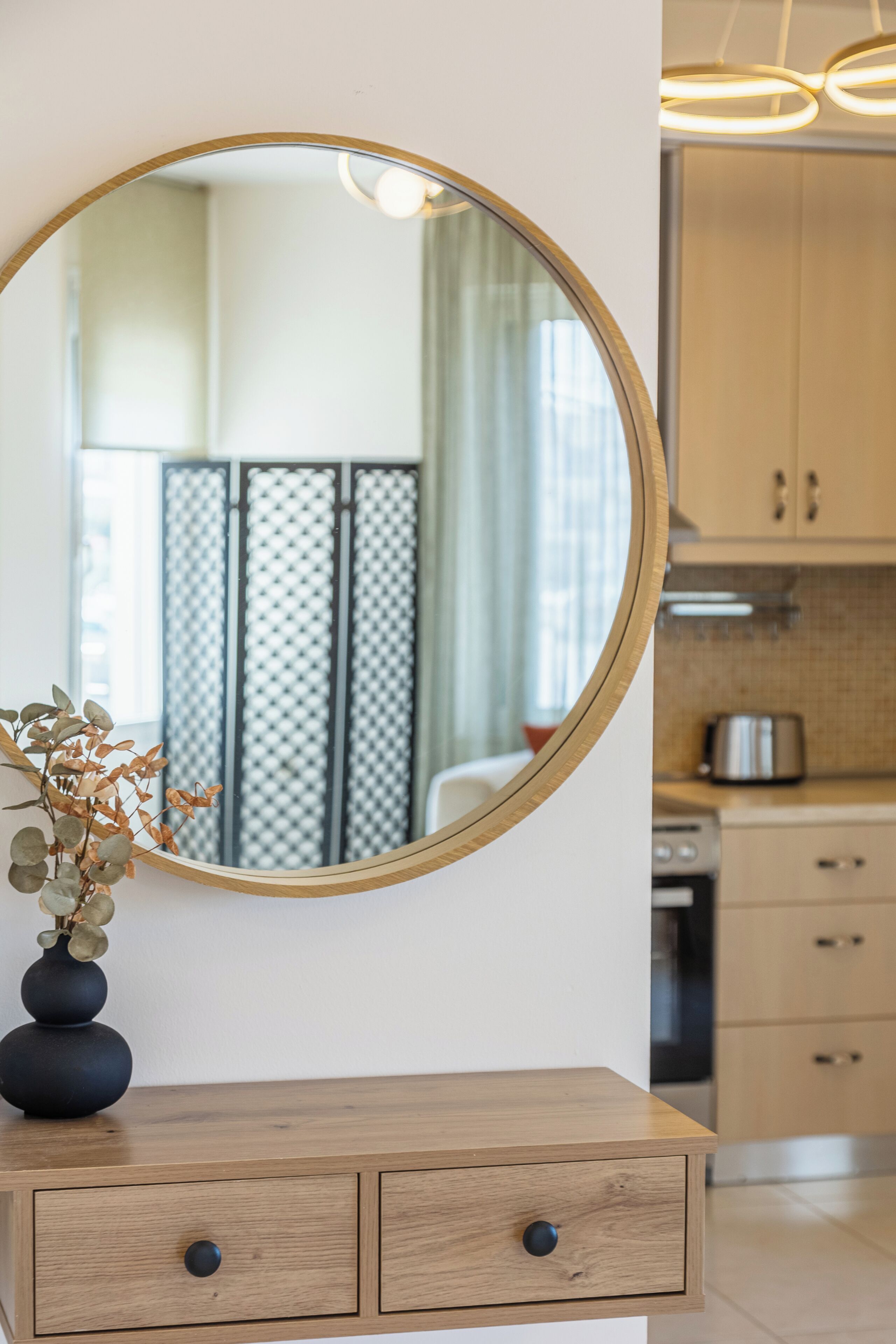 Een ronde spiegel op een houten dressoir, met een vaas gedroogde bloemen. Op de achtergrond een keuken met kastjes en apparatuur.