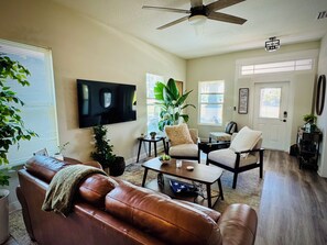 A living room with a brown leather sofa, a coffee table, a ceiling fan, a TV mounted on the wall, and a door with a window. - Charming 3-bedroom house in Ybor/Tampa! Walk to Trolley for events (Tampa)