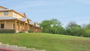 A multi-story residential building with a covered porch, a grassy hill, and trees in the background. - New Braunfels | Friends River Escape | Pool + River | 2BR (New Braunfels)