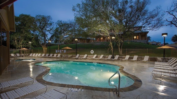 A pool area with lounge chairs, a hot tub, and a building in the background. - New Braunfels | Friends River Escape | Pool + River | 2BR (New Braunfels)