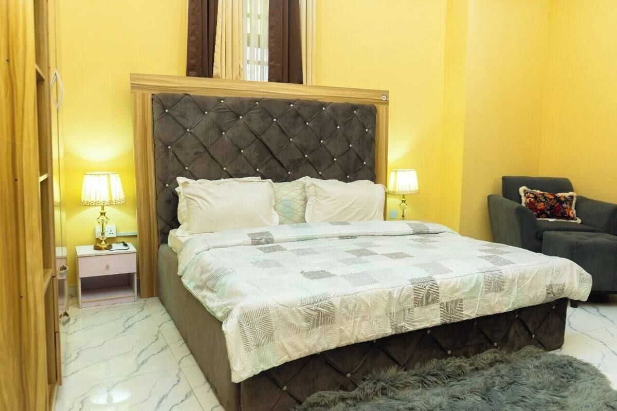 A bedroom with a tufted headboard, a bed with a patterned bedspread, bedside lamps, a nightstand, a chair, and a fluffy rug.