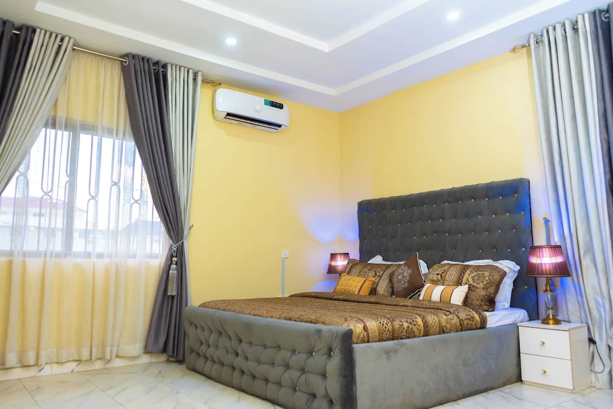 A bedroom with a large bed, an air conditioning unit, a bedside lamp, and curtains.