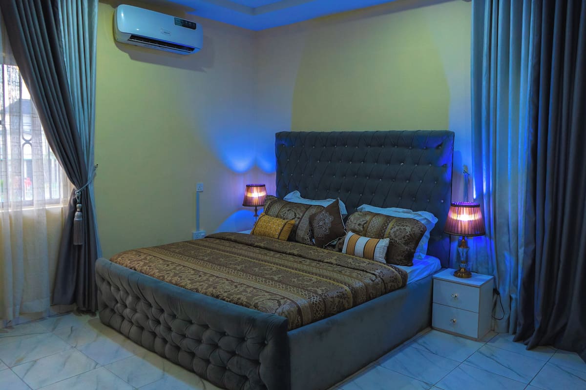 A bedroom with a tufted headboard, a bed with patterned bedding, two bedside lamps, a nightstand, and an air conditioning unit.