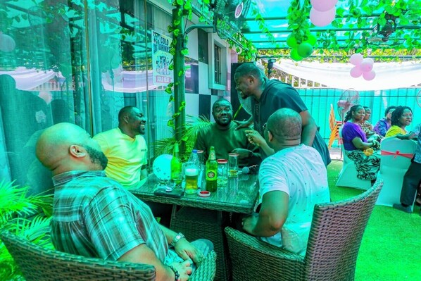 A group of people seated at an outdoor table with drinks and a fan, surrounded by greenery and a checkered wall. - 114 Luxury Apartments by Oragon Hotel (Lagos)