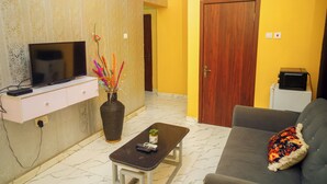 A living room with a television on a stand, a small table with a remote control, a sofa with a decorative pillow, and a door in the background. - 114 Luxury Apartments by Oragon Hotel (Lagos)