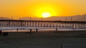 Une plage avec une jetée qui s’avance dans l’eau, des gens qui se promènent le long du rivage et un coucher de soleil en arrière-plan.