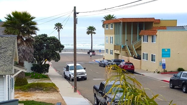 Vista di una strada con un edificio dotato di una scala che conduce a un piano superiore, un parcheggio con diverse auto e vista sull'oceano.