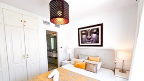 Habitación de hotel con cama, cabecero, mesita de noche, lámpara, armario y baño visible a través de una puerta abierta.