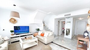 Una sala de estar moderna con un televisor de pantalla plana, sofás blancos, una mesa de centro y una escalera que lleva a otra habitación.