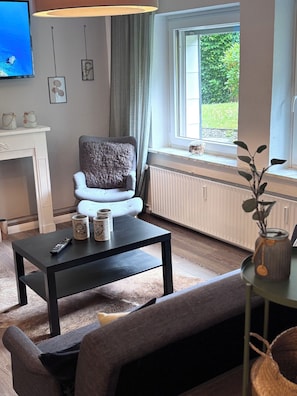 Ein Wohnzimmer mit Kamin, einer Couch, einem Couchtisch mit Fernbedienung und einem Fenster mit Blick ins Grüne.