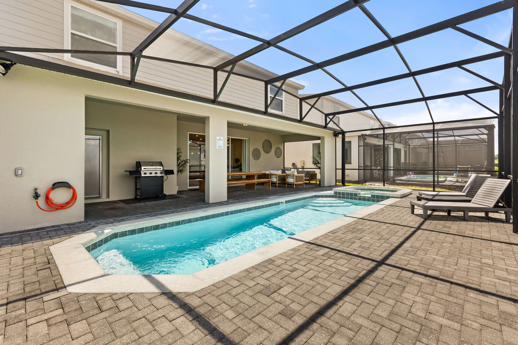 A pool area with a covered patio, lounge chairs, and a grill.