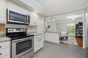 A modern kitchen with white cabinetry, a built-in microwave, a stove, and an oven. There is a dining area with a table and chairs, and a bookshelf filled with books. - WinStar Getaway: 3 bedroom 2 full bath - Sleeps 6-10 - Lot of Parking - EV Port (Gainesville)