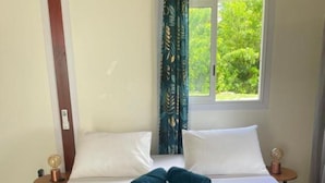 Una cama con almohadas blancas y una colcha azul oscuro, una ventana con vistas a follaje, y un techo de madera.
