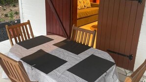 Mesa al aire libre con mantel estampado, sillas de madera y vista a una habitación con cama y puerta.