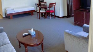 Una habitación de hotel con una cama, una mesita con un control remoto, un sofá, una mesa de comedor con sillas, un mueble para televisión y un aire acondicionado.