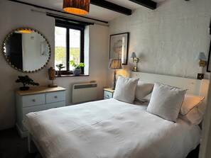 A bedroom with a bed, bedside tables, a dresser, a radiator, a window with plants, a mirror, and wall-mounted lamps. - Smugglers Cottage Barn Conversion with Indoor Pool 
Jurassic Coast AONB (Whitchurch Canonicorum)