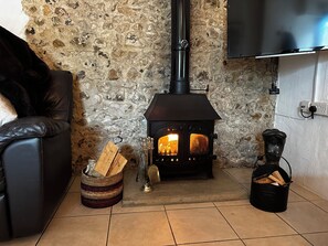 A cozy living room with a stone wall, a black leather sofa, a fireplace with logs, and a television mounted on the wall. - Smugglers Cottage Barn Conversion with Indoor Pool 
Jurassic Coast AONB (Whitchurch Canonicorum)