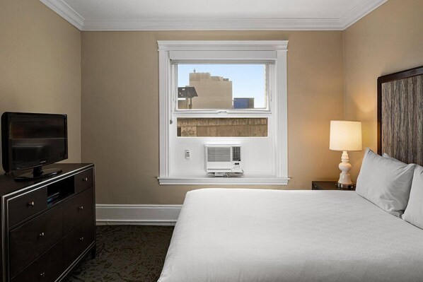 A hotel room with a bed, a dresser, a television, a window with an air conditioner, and a lamp.