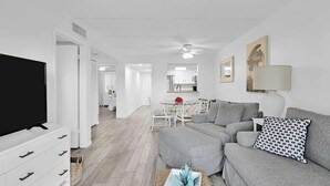 A modern living room with a flat-screen TV, grey sofa, and a dining area with a glass table. - Beachside Condo Resort Sleeps 6 in Beautiful St Augustine! (St. Augustine)