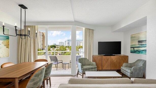 A modern hotel room with a dining area, seating, a flat-screen TV, and a balcony with a city view. - Santa Barbara | Romantic Beach Retreat | Pool + Beach (Pompano Beach)