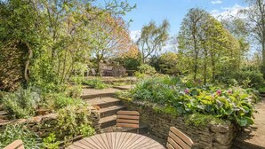 A garden with a circular wooden table and benches, surrounded by various plants and a stone wall. - Cottage Sleeps 6 With Parking and Garden (Shipston on Stour)