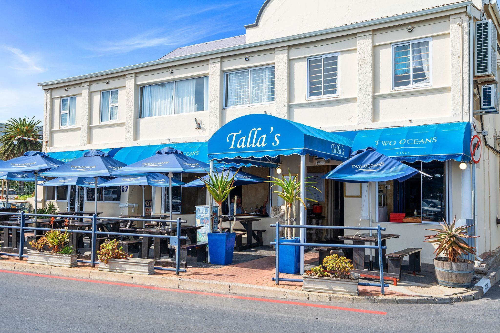 A seaside restaurant with outdoor seating, blue awnings, and potted plants.