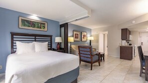 Una habitación de hotel con cama, una silla, un escritorio, kitchenette y cuadros en la pared.