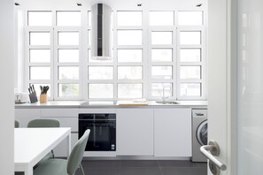 A modern kitchen with white cabinetry, a built-in oven, a dishwasher, and a window with multiple panes. - Master Deco Gem Avenidas Novas I – GEM Residence (Lisbon)