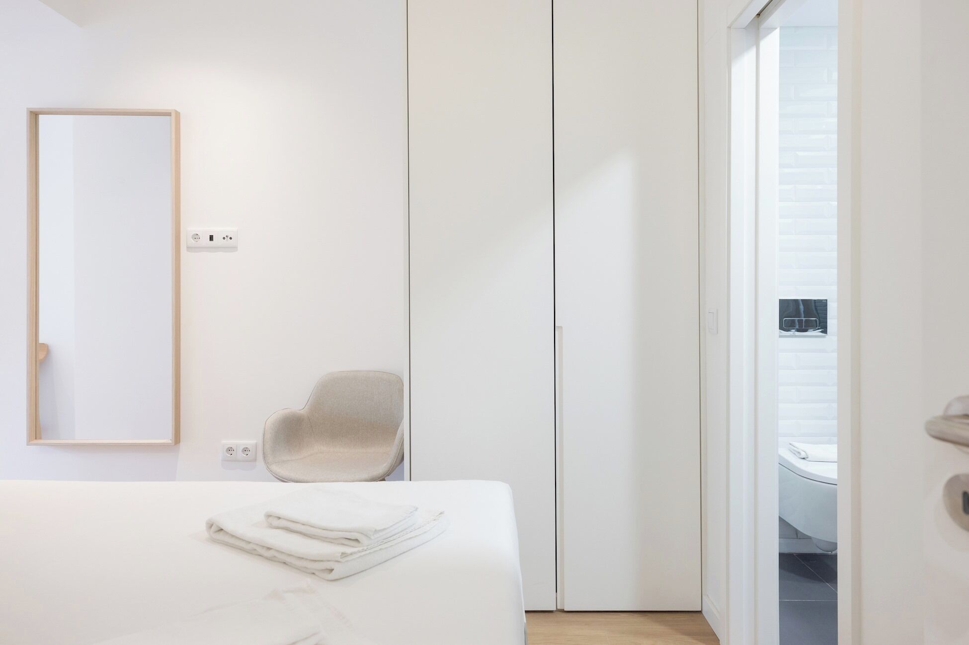 Ein schlicht eingerichtetes Schlafzimmer mit einem Bett, einem Sessel, einem Spiegel und einer Tür zu einem Badezimmer.