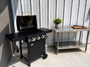 Una parrilla negra portátil sobre ruedas, una mesa metálica con una planta en maceta y una tabla de cortar de madera.