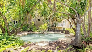 A natural pool surrounded by lush greenery and trees, with a building in the background.