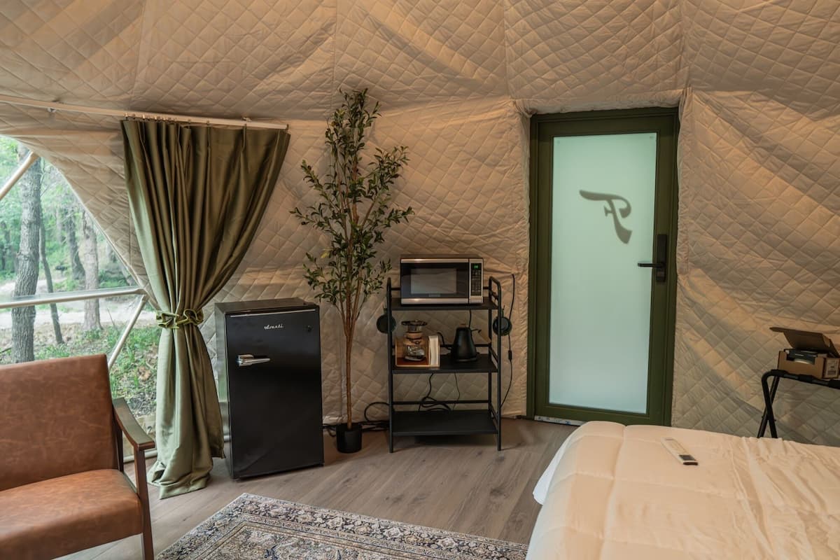 Una habitación con carpa, una cama, un escritorio pequeño con microondas, una estantería y una puerta con un símbolo.