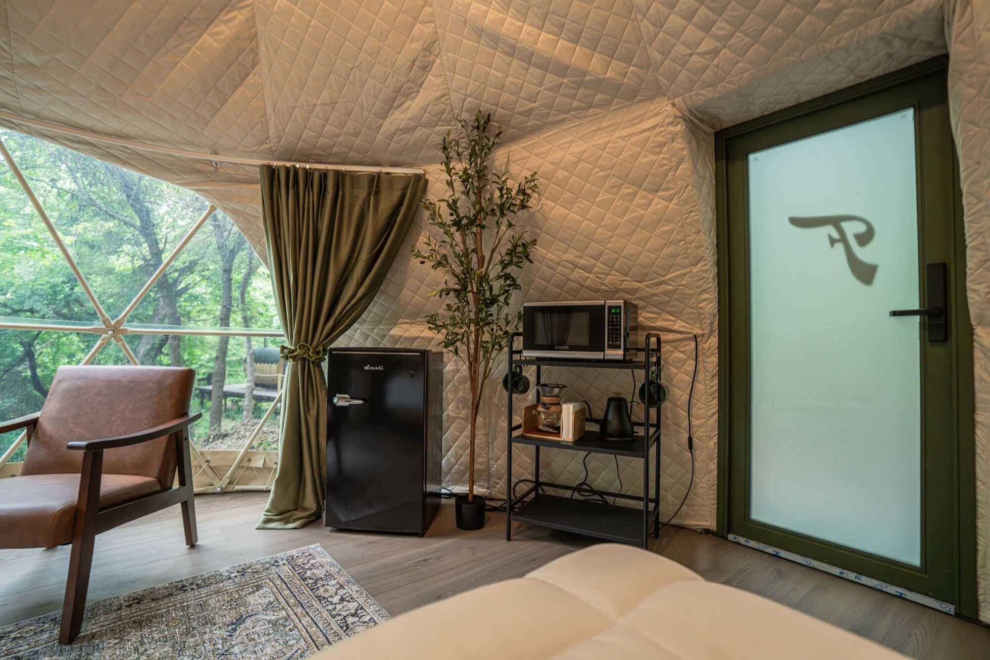 A cozy tented room with a bed, a chair, a small fridge, a microwave, and a shelf with various items.