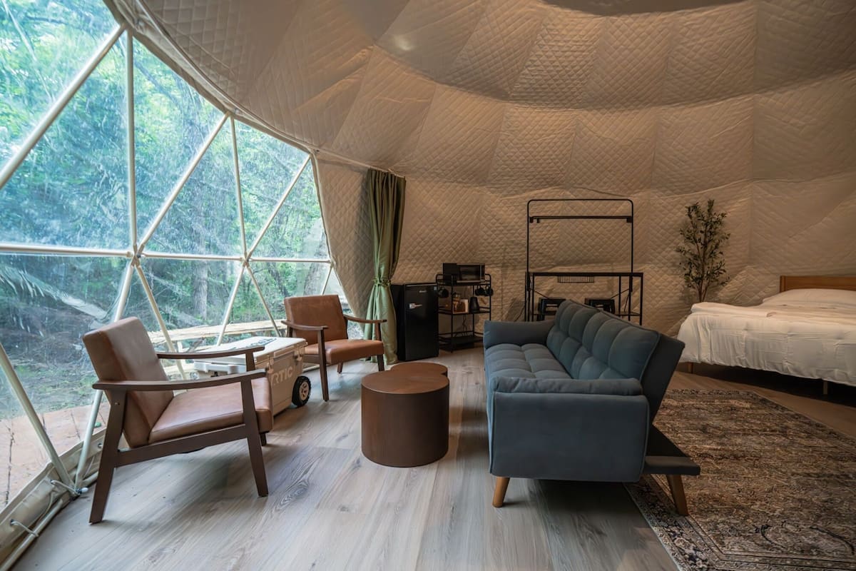 Un interior acogedor con una cama, sofá, sillón y una mesita, todo dispuesto alrededor de un ventanal con vistas al bosque.