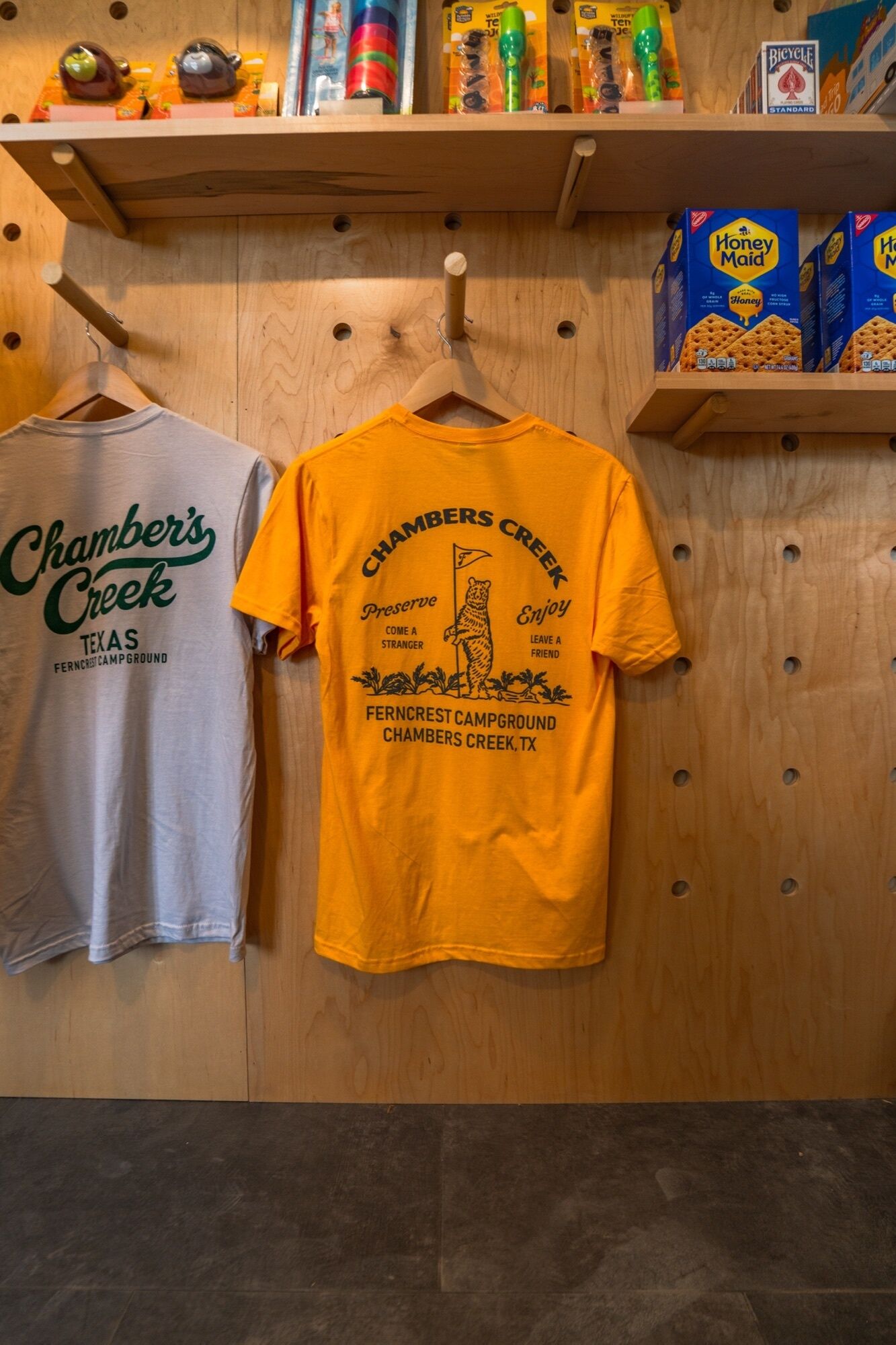 Twee t-shirts hangen aan een houten wand, waarvan er een 'Chamber's Creek Texas' en de andere 'Chambers Creek Preserve' zegt.