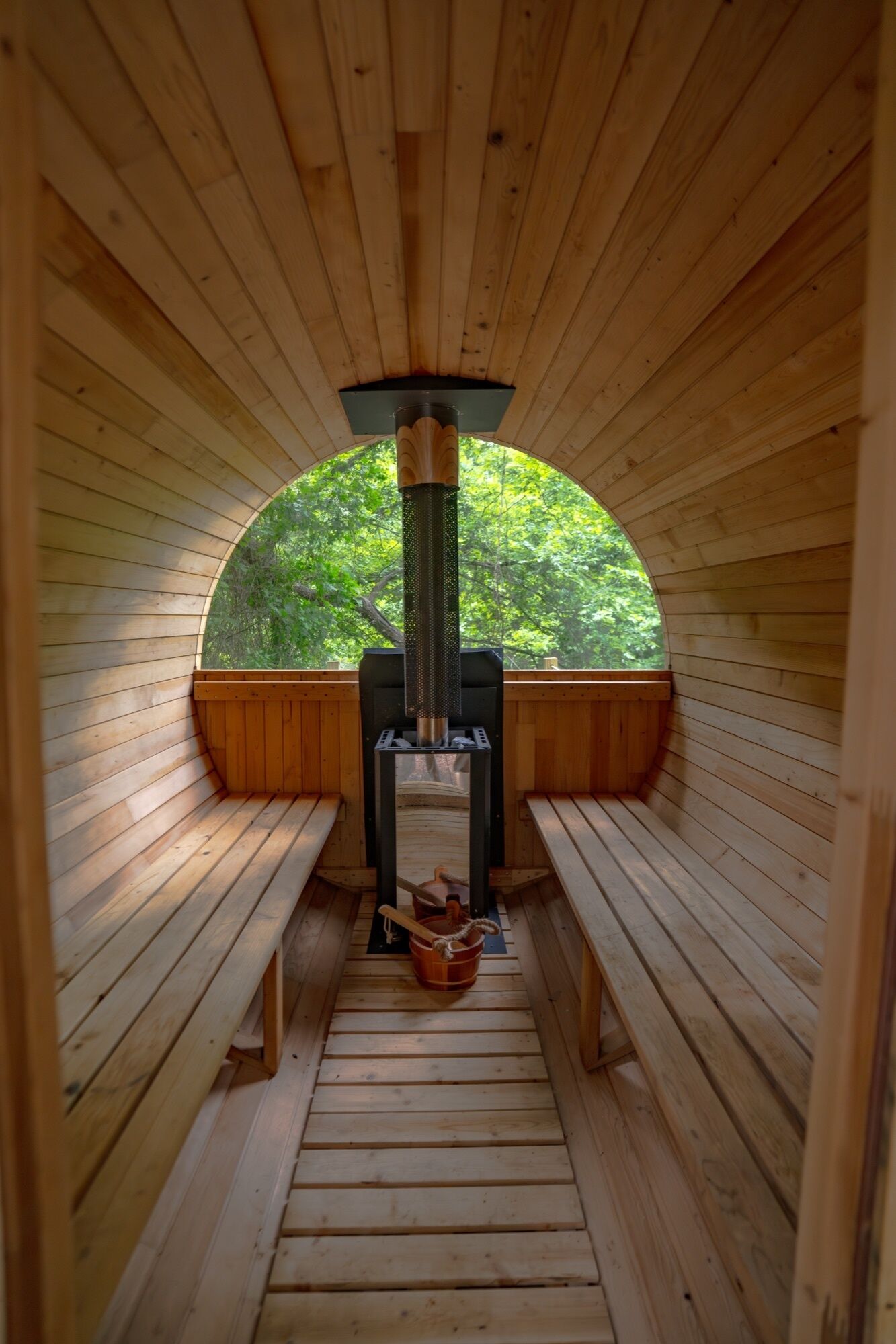 Un sauna avec des bancs en bois, un radiateur central et une vue sur la verdure grâce à une ouverture en demi-cercle.