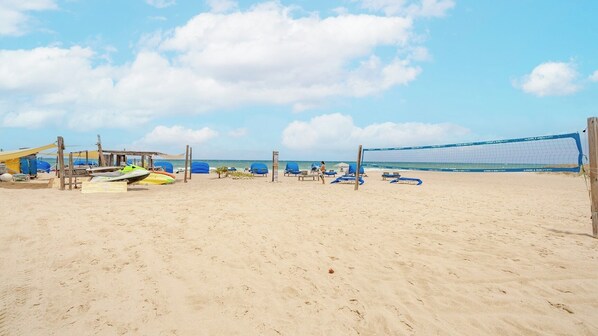 Ein Strand mit einem Volleyballnetz, Liegestühlen und einer kleinen Konstruktion mit Tisch.