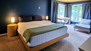 Ett sovrum med en säng, två sänglampor, en takfläkt, ett fönster med gardiner och ett litet bord med en stol.