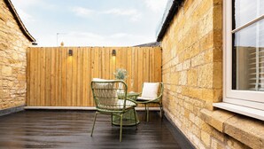 Eine Holzterrasse mit zwei Korbstühlen und einem Tisch, eine Steinmauer und ein Fenster mit Jalousien.