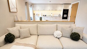 Ein modernes Wohnzimmer mit einem beigefarbenen Sofa, einem Couchtisch und einer Küche im Hintergrund.