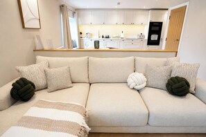 A modern living room with a beige sofa, a coffee table, and a kitchen in the background. - Lygon Lookout | Cotswolds Stay for 2 | Pet-friendly w/ Roof Terrace (Worcestershire)