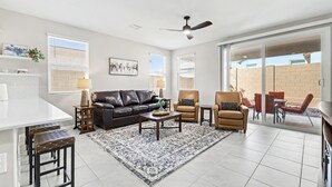 Una sala moderna con un sofá de cuero, dos sillones, una mesa de centro y un tapete estampado. Cuenta con un ventilador de techo, un comedor con mesa y sillas, y un amplio ventanal corredero que da a un patio exterior.