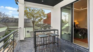 Un balcon avec une table et des chaises, une porte vitrée donnant sur une chambre, et des panneaux solaires sur le toit d’un immeuble voisin.