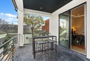 Ein Balkon mit Tisch und Stühlen, eine Glastür, die zu einem Schlafzimmer führt, und Solaranlagen auf einem Nachbardach.