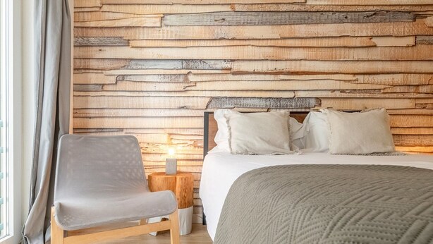 Un dormitorio con pared de madera, una cama con ropa blanca, una silla gris y una mesita de noche con una lámpara.