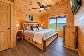 Ein Schlafzimmer einer Holzhütte mit einem Bett, einem Nachttisch, einem Deckenventilator, einem Fenster mit Blick und einem Wand-Fernseher.