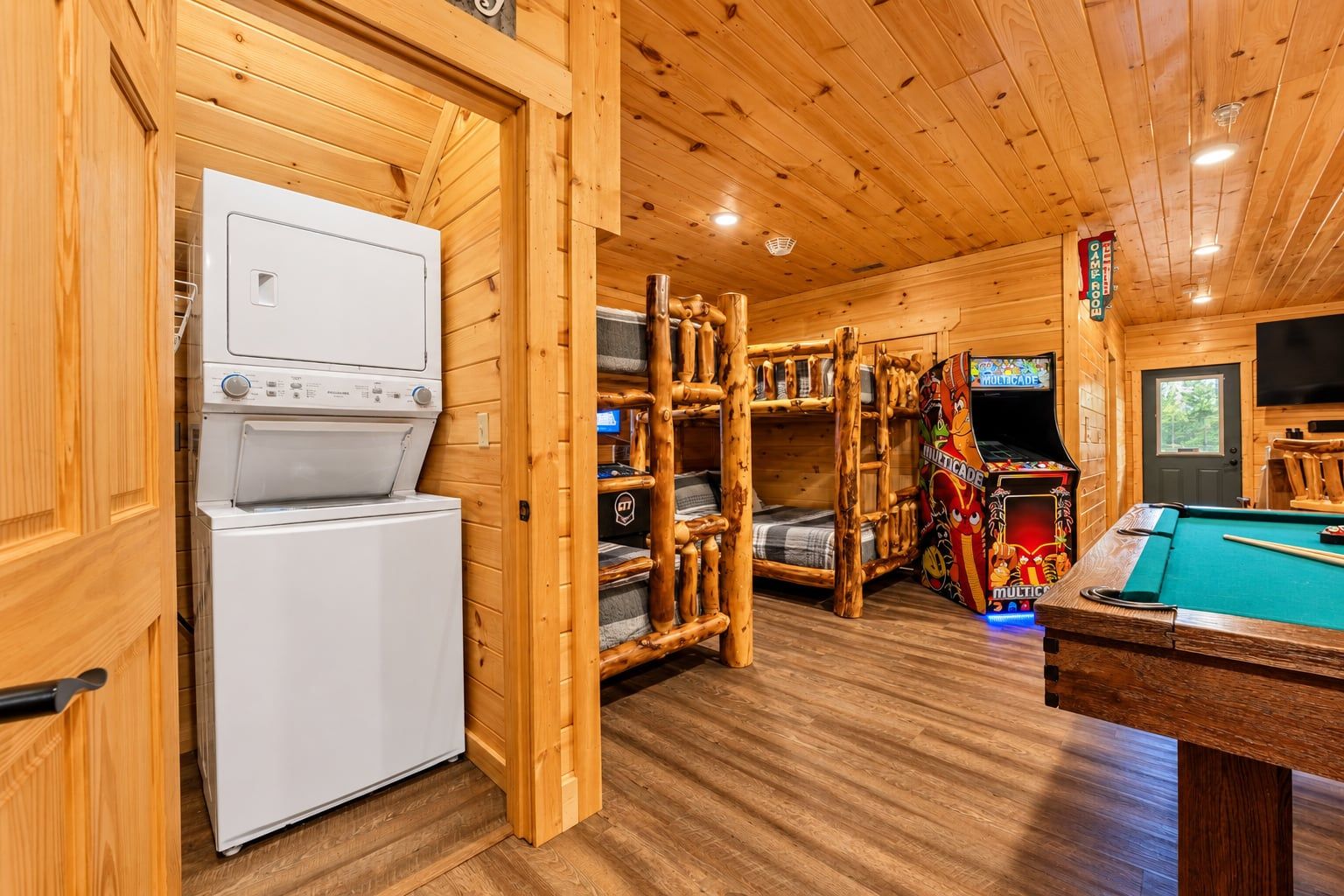 A wooden cabin interior with a pool table, bunk beds, a washing machine, and a television.
