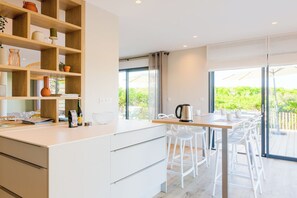 A modern kitchen with white cabinetry, a wooden shelf with decorative items, a dining area with white chairs, and a large window with a view of greenery. - Apartment 50 metres from the sea (Penmarch)