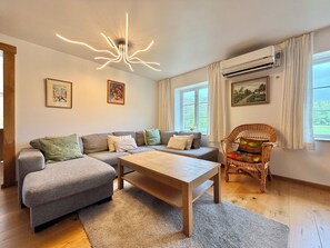 Een woonkamer met een grijze bank, een houten bijzettafeltje, een rieten fauteuil en een plafondlamp.
