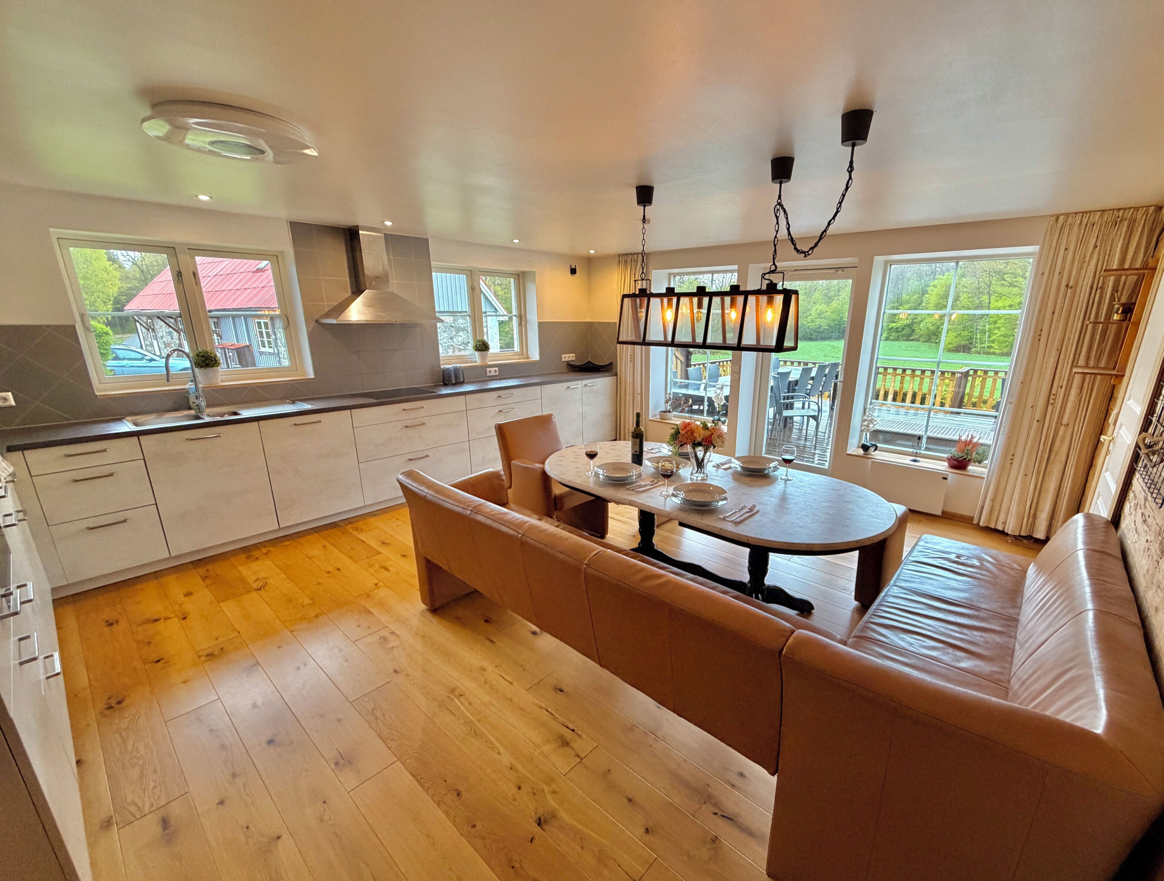 A modern kitchen with a central island, wooden flooring, and large windows offering views of the outdoors.